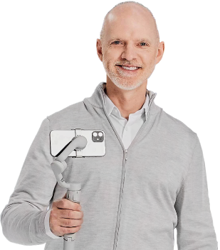Man wearing a gray sweater holding a camera on a selfie stick with no backgroud.